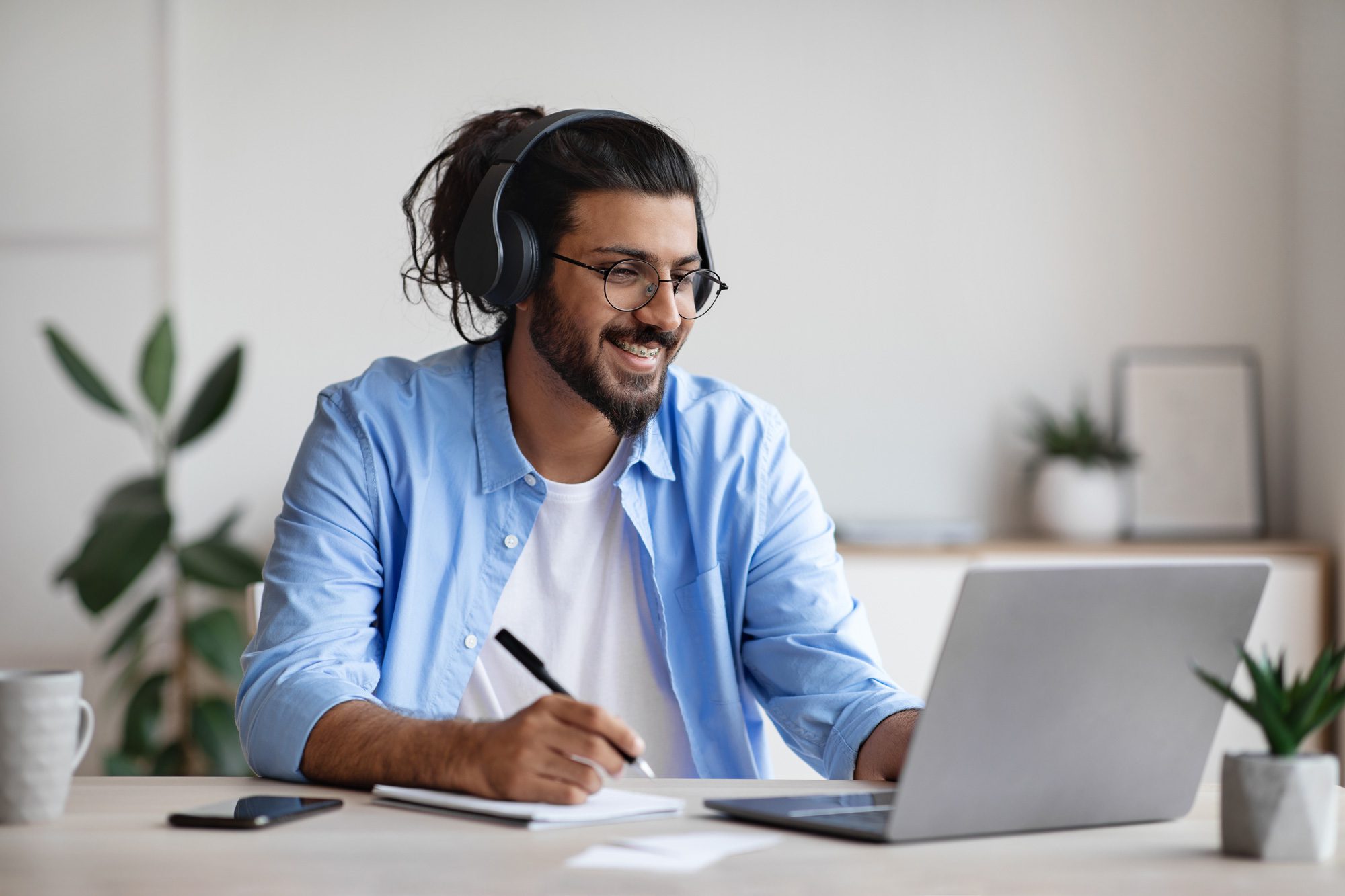 smiling man wearing headphones while looking at a computer and writing. university of Dubuque online admissions